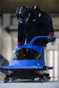 Macy Tarlton is in a black bobsled suit and sprinting and pushing a blue bobsled on the track. 