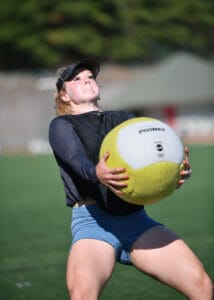 Macy Tarlton is bracing while holding a large yellow medicine ball and is about to throw it in the air. 